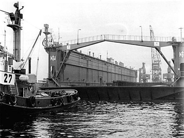 Das Schwarz-Weiß-Bild zeigt ein Schwimmdock der Warnowwerft im Überseehafen Rostock. Davor fährt ein Schlepper.