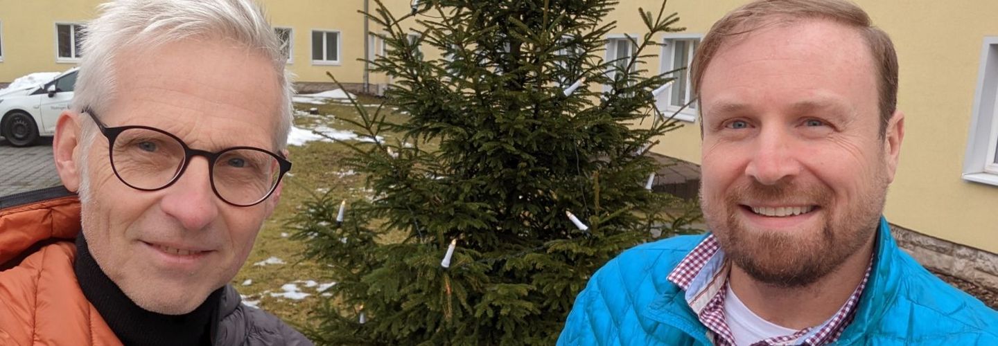 Maximilian Schönherr und Sascha Münzel stehen vor einem Weihnachtsbaum, der mit Lichtern geschmückt ist.