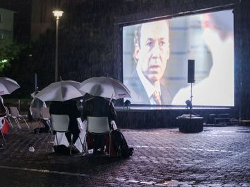 Zuschauer unter Regenschirmen sitzen vor der Kinoleinwand. Regentropfen prasseln herab.