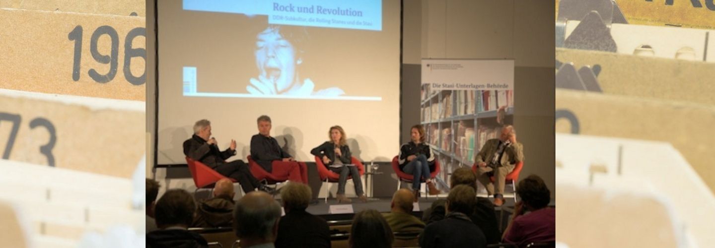 Auf einer Bühne sitzen vier Personen in roten Sesseln. Im Hintergrund ist auf einer Leinwand das Cover des Dokumentenhefts "Rock und Revolution" zu sehen.