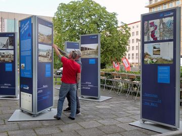 Blaue Schautafeln mit vielen Fotos der Ausstellung "Das Verschwinden der Mauer". Vor einer der Schautafeln stehen zwei Besucher, die auf eines der Fotos zeigen.