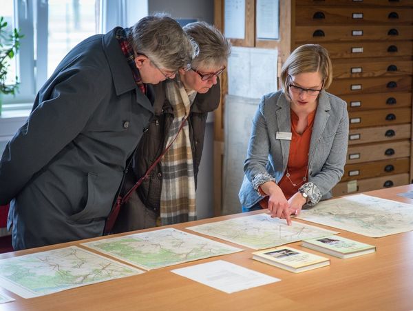 Eine Mitarbeiterin des Bundesarchivs zeigt einer Besucherin und einem Besucher Karten aus den Beständen des Bundesarchivs
