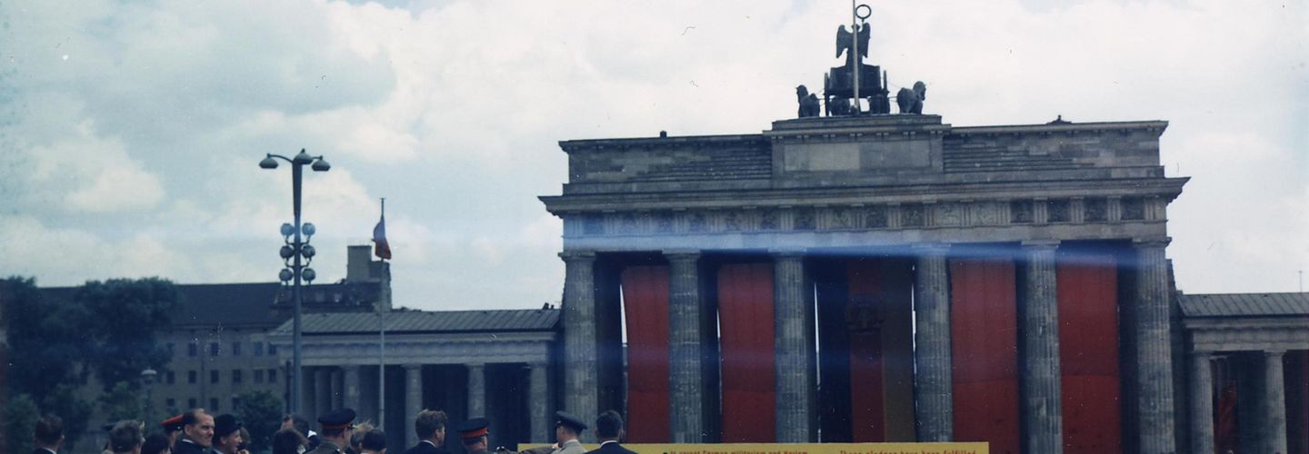 US-Präsident John F. Kennedy steht auf einem Podest und blickt auf das mit roten Tüchern verhängte Brandenburger Tor.