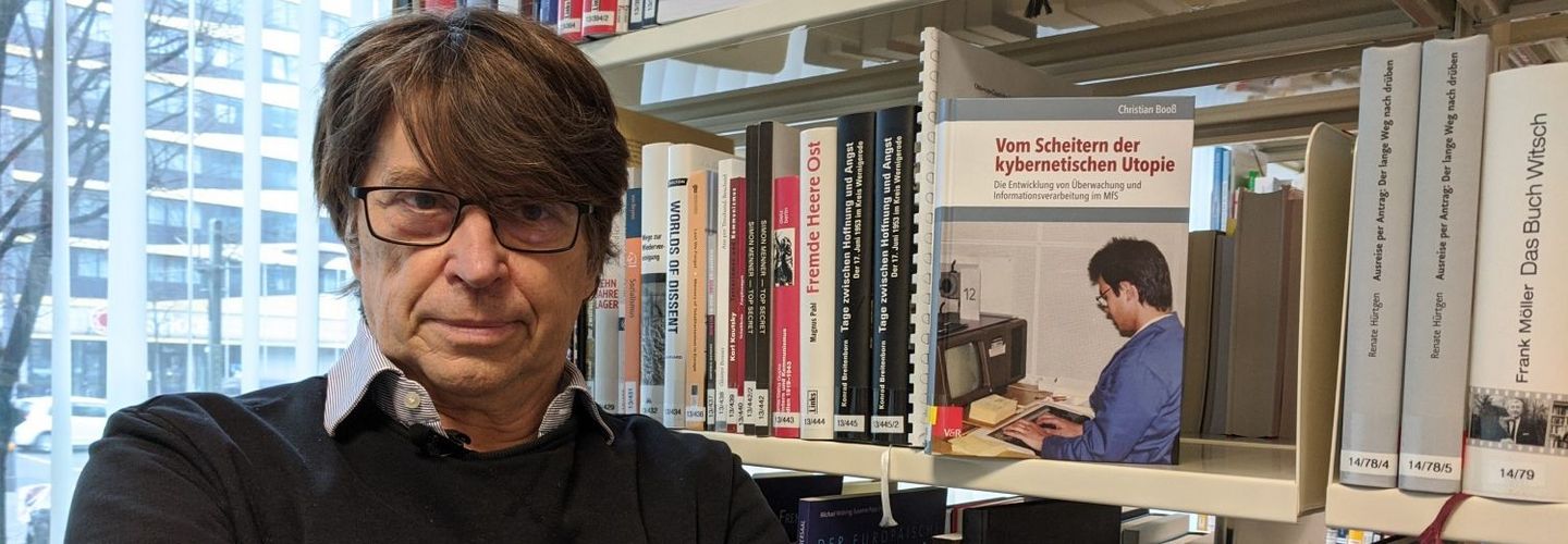 Dr. Christian Booß steht neben einem Bücherregal, in dem sein Buch Dr. Christian Booß neben seiner Publikation "Das Scheitern der kybernetischen Utopie" zu erkennen ist.