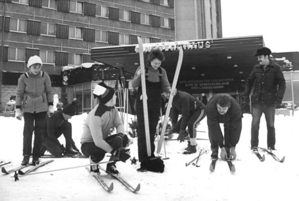 Jugendliche in Winterbekleidung beim Anschnallen ihrer Skier vor dem Ringberghaus in Suhl. Über dem Eingang des Gebäudes steht in Versalien: "Wir grüssen die Teilnehmer zum 26. Winterurlaub der Landjugend".
