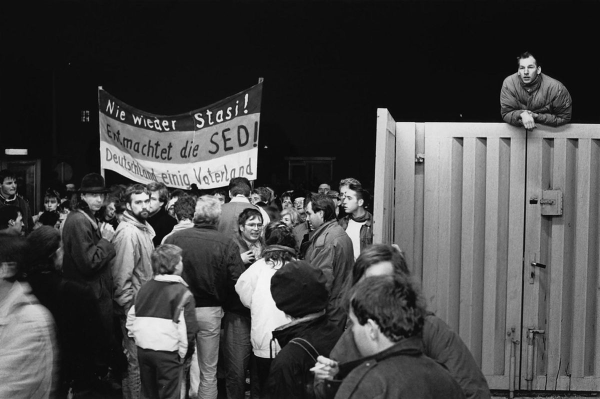 Das Schwarz-Weiß-Foto zeigt eine Gruppe von Menschen am geöffneten Haupteingang der Stasi-Zentrale an der Ruschestraße/Normannenstraße. Auf einem Banner steht "Nie wieder Stasi! Entmachtet die SED! Deutschland einig Vaterland". Oben rechts im Bild lehnt sich ein Mann über das Eingangstor.  