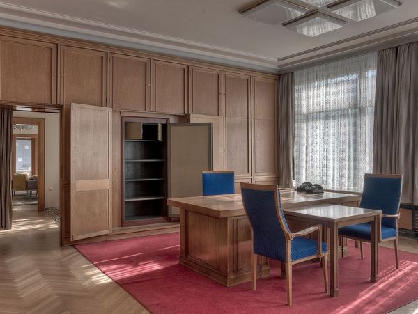 Das Bild zeigt ein hochwertig eingerichtetes Büro mit Holzverkleidung. In der Mitte des Raumes steht ein großer Schreibtsich mit drei Stühlen. Die Fenster sind mit halbdurchlässigen Vorhängen abgedeckt.