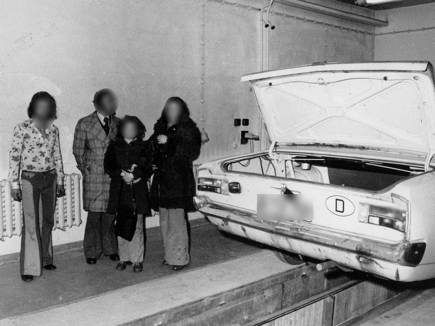 Das Bild zeigt ein Westberliner Auto, das Stasi-Mitarbeiter von hinten fotografierten. Das Fahrzeug steht ebenerdig auf einer Art Hebebühne. Der Kofferraum ist geöffnet. Links vom Fahrzeug stehen vier Personen. Ganz links der vermeintliche Fahrer. Daneben die drei Personen, die sich im Kofferraum versteckt hatten.