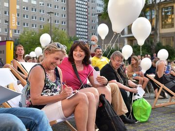 Zwei Besucherinnen füllen lachend einen Zettel während der Ballonaktion aus. In den Händen halten sie einen weißen Ballon.