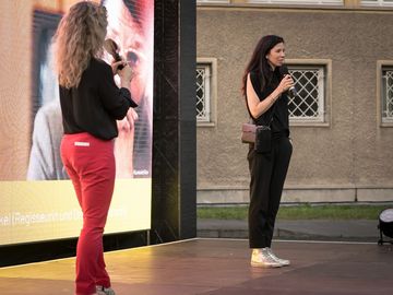 Auf einer Bühne stehen vor der Filmleinwand zwei Frauen. Links ist Dagmar Hovestädt, die Moderatorin zu erkennen. Rechts spricht Regisseurin Franziska Stünkel in ein Mikrofon.