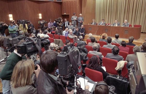 Zu sehen ist Günter Schabowski auf dem Podium. Im Vordergrund befinden sich teils stehende, teils sitzende Presseleute, die sich dem Podium zuwenden.