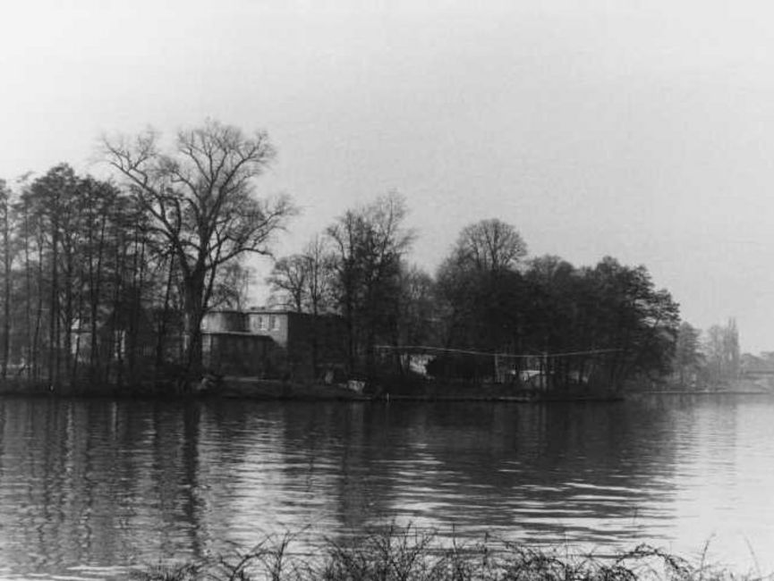 Aufgenommen vom gegenüberliegenden Ufer des Gewässers ist das Haus kaum zwischen den zahlreichen, überwiegend kahlen Bäumen auszumachen. Es handelt sich um ein schwarz-weißes Lichtbild.