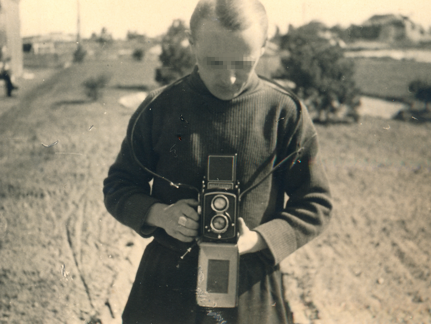 Das schwarz-weiße Lichtbild zeigt einen blonden Fotografen, die Frontalaufnahme geht bis über die Hüfte. Er blickt nach unten auf seine Kamera, im Hintergrund erstreckt sich eine weite Landschaft mit wenigen kleinen Bäumen.