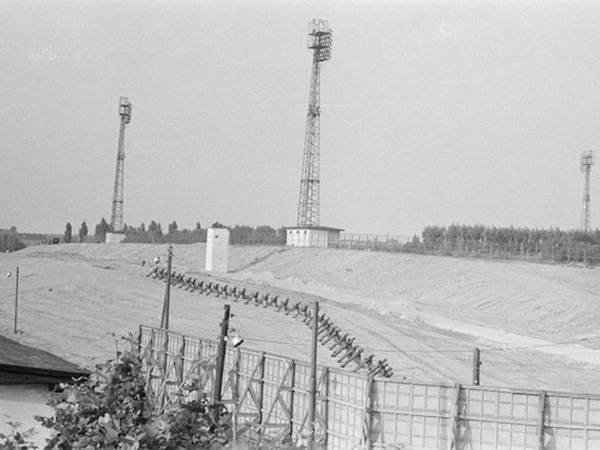 Das schwarz-weiße Lichtbild zeigt die Sicherungsanlagen der innerdeutschen Grenze. Auf einem Hügel stehen im Hintergrund drei Funktürme, die umzäunt sind.