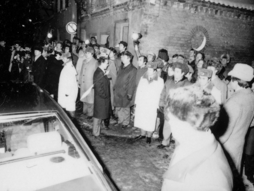 Die Schwarzweißaufnahme zeigt Zuschauer und Presse am Straßenrand bei Helmut Schmidts Staatsbesuch in der DDR 1981. Die Leute von der Presse richten ihre Kamera auf die Straße, auf der ein fahrendes Auto am linken Bildrand erkennbar ist.  