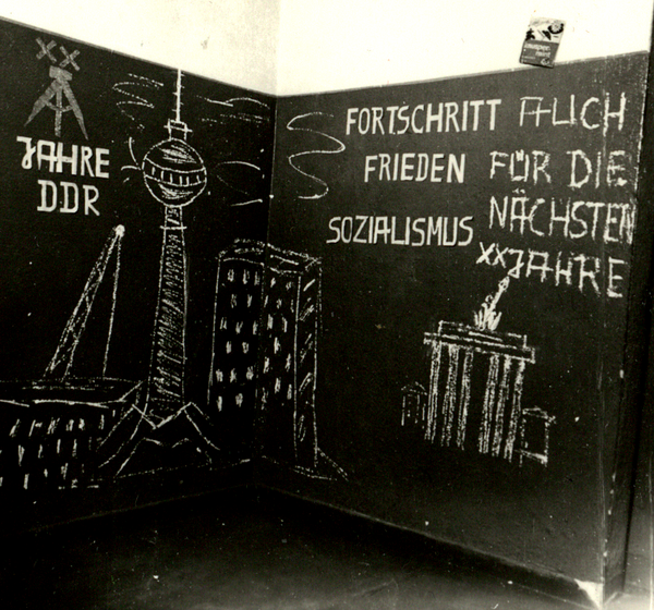 Auf einer Tafel steht mit Kreideschrift "Fortschritt Frieden Sozialismus auch für die nächsten XX Jahre". Daneben sind der Berliner Fernsehturm und das Brandenburger Tor abgebildet.