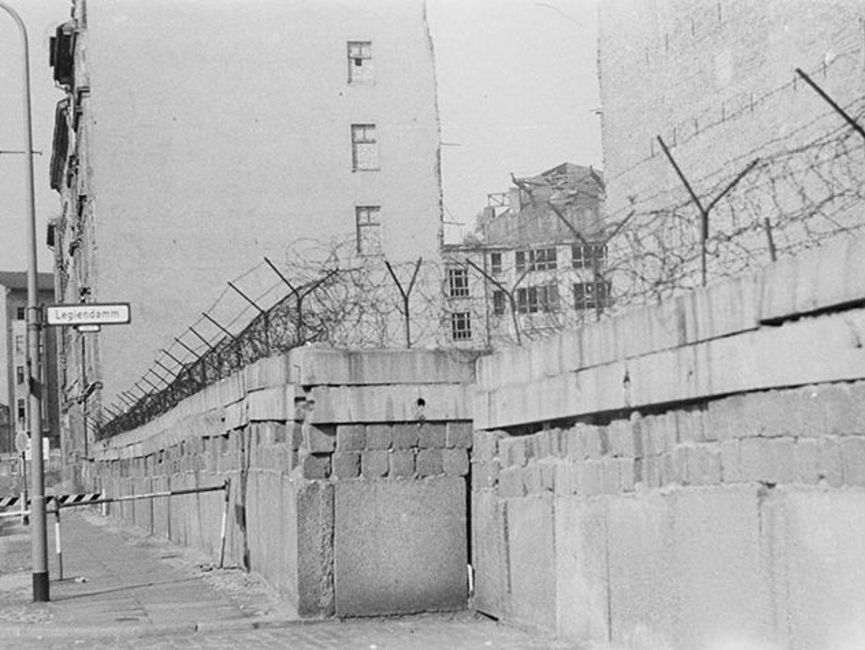 Abgebildet ist der Blick auf die Berliner Mauer vom Westteil der statt. Auf einem Straßenschild ist deutlich "Legiendamm" zu lesen.