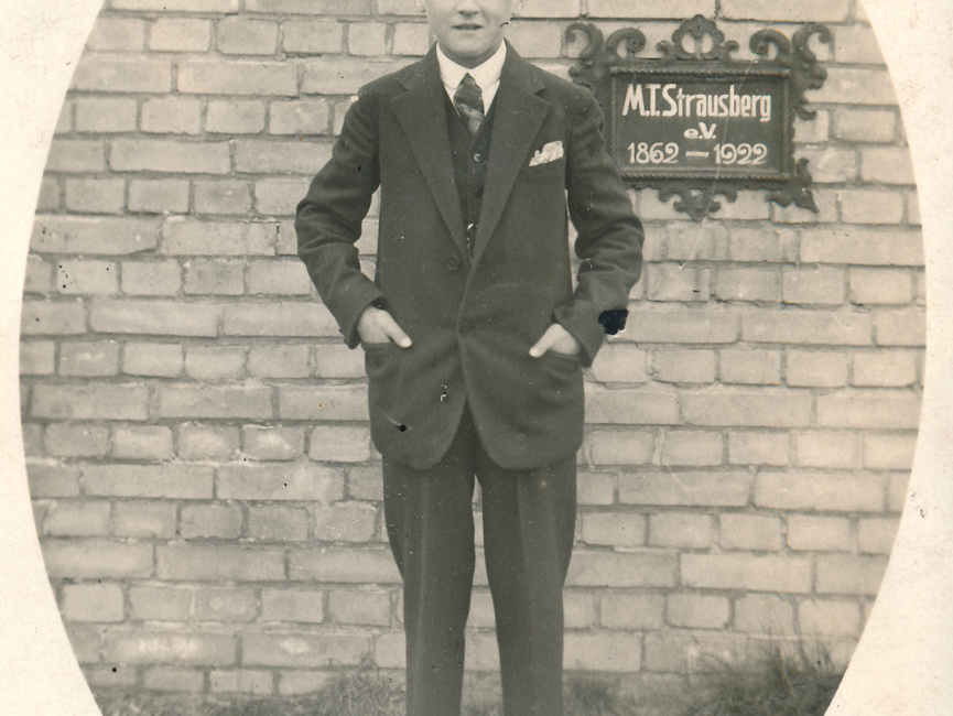 Das ovale Lichtbild ist schwarz-weiß und zeigt einen Jungen im Dreiteiler Anzug. Er steckt die Hände in die Hosentasche, blickt frontal in die Kamera. Hinter ihm steht eine Ziegelmauer, auf der ein Schild "M. T. Strausberg e.V.; 1962-1922" angebracht ist.