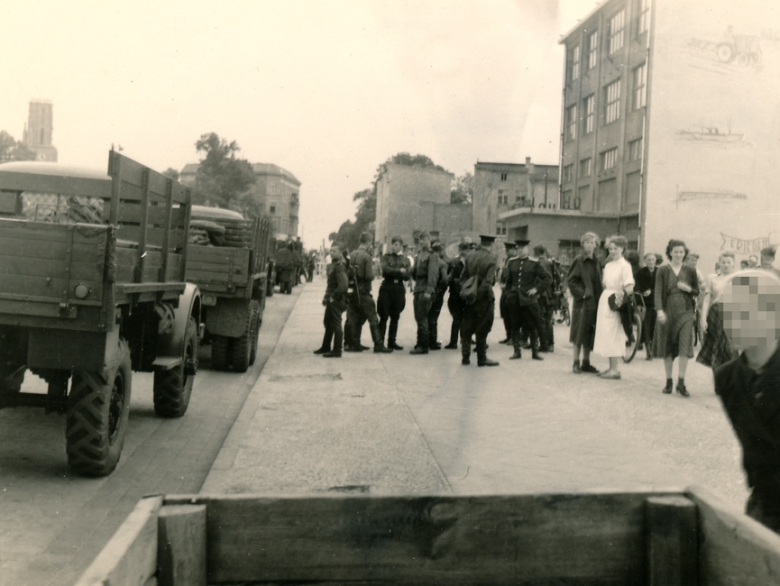 Diese Aufzeichnung zeigt sowjetische Soldaten, welche "Das Haus der Freundschaft" in Rathenow sichern. Auf der linken Seite der Aufzeichnung befindet sich eine Straße auf der vier sowjetische Truppentransportfahrzeuge parken und auf der rechten Seite befinden sich ein paar Zivilisten.