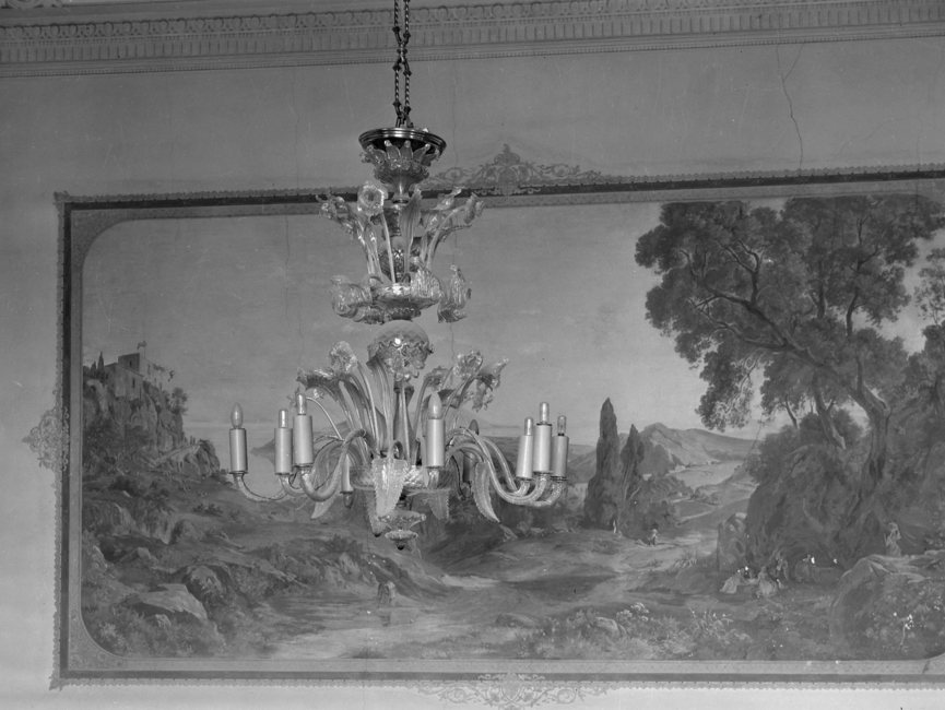 Auf dem schwarz-weißen Lichtbild ist ein Kronleuchter zu sehen, der vor einem gemalten Landschaftsbild hängt. Am Deckenübergang ist Stuck angebracht.