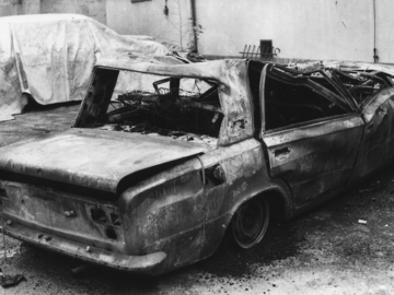 Das Schwarz-Weiß-Bild zeigt ein völlig ausgebranntes Fahrzeug. Links davon steht ein weiteres Fahrzeug, das mit einer Plane abgedeckt wurde.