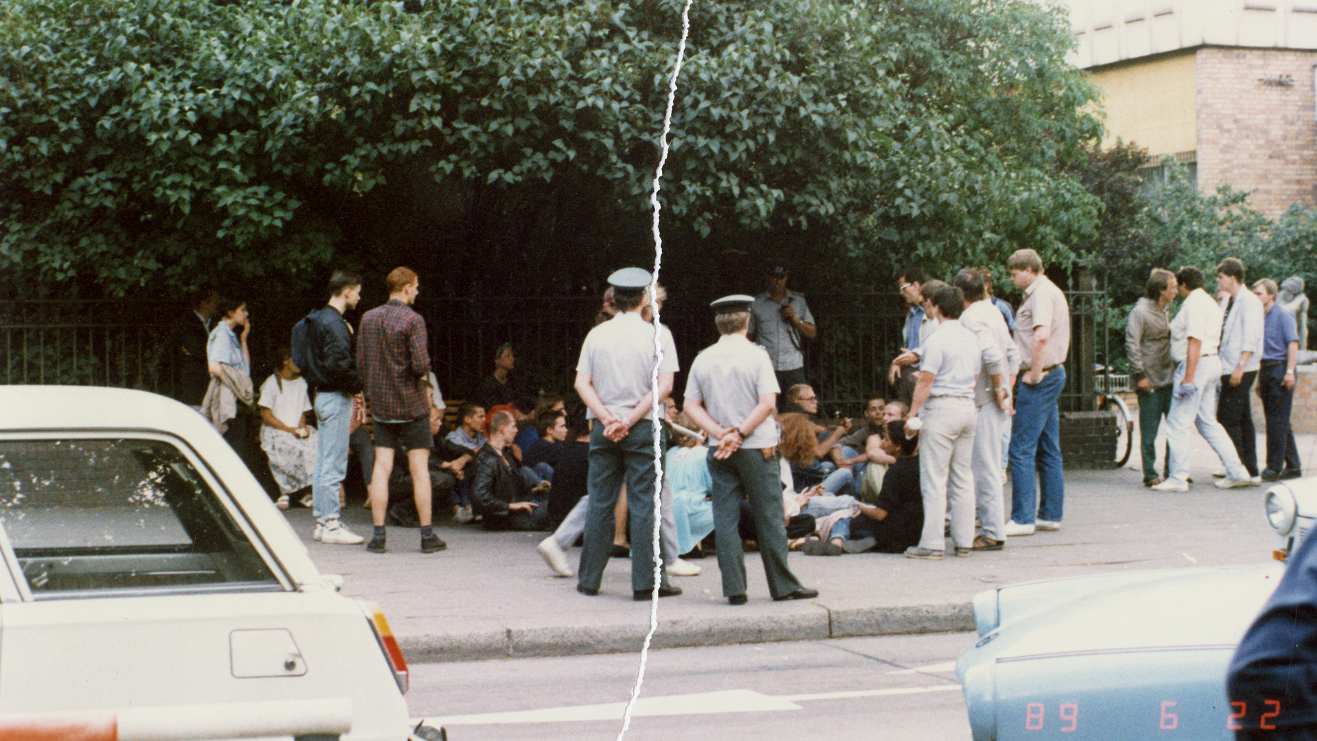 Eine Personengruppe sitzt auf einem Bürgersteig direkt vor einem Zaun. Sie werden umringt von Volkspolizisten sowie einigen steif wirkenden Herren in ordentlichem Zivil. Das farbige Lichtbild wurde manuell rekonstruiert, in der Mitte ist der vertikale Riss sichtbar.