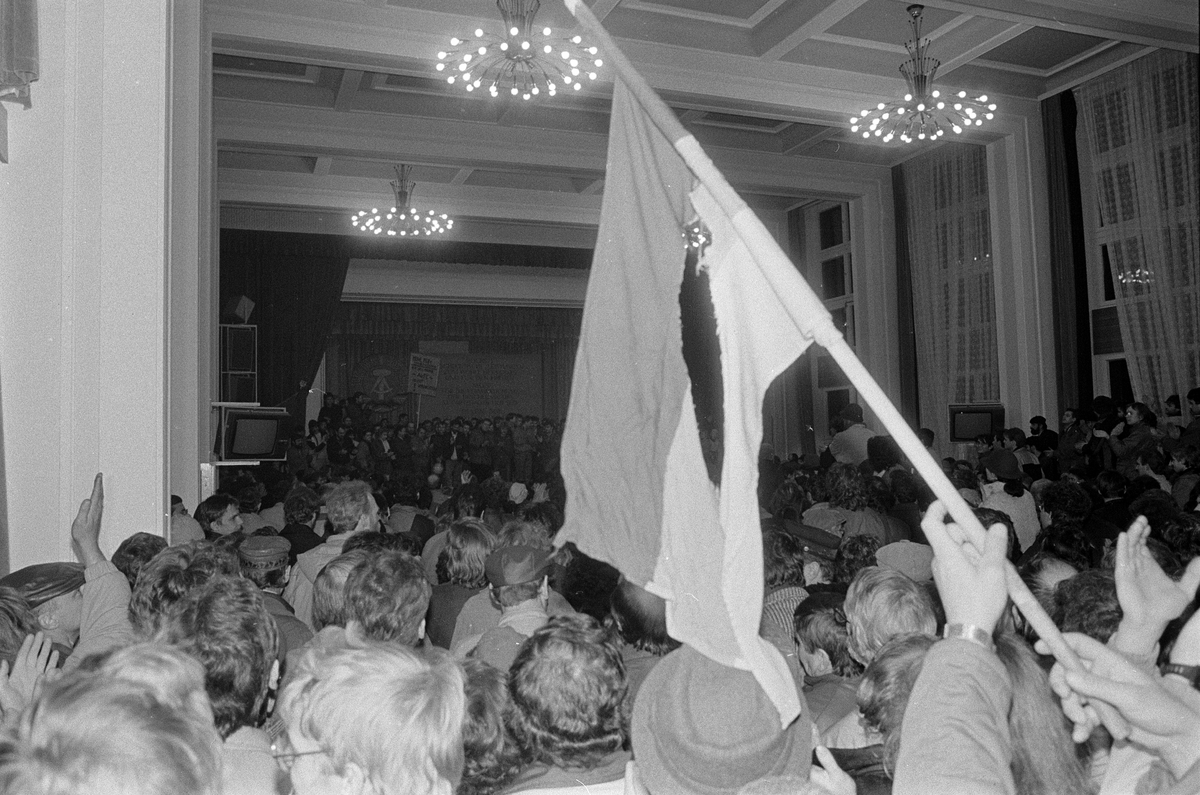 Das Bild zeigt eine große Menschenmenge in einem großen Saal. Im Vordergrund schwenkt jemand eine nicht genau zu identifizierende Fahne.