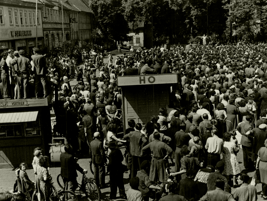 Noch einmal der Marktplatz des thüringischen Sömmerda am 17. Juni 1953. Im Hintergrund die Menschenmenge, die anscheinend einer Kundgebung beiwohnt. Im Vordergrund die Bretterbuden "Zeitungsvertrieb" und "HO, auf denen Menschen sitzen oder stehen. Ganz vorne weitere Menschen, manche von ihnen mit Fahrrädern. 