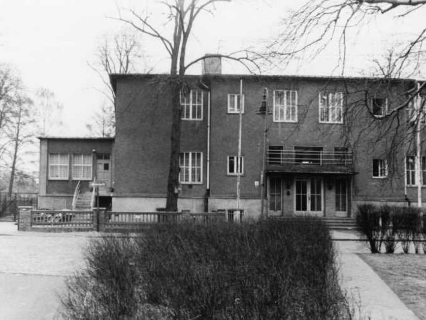 Das schwarz-weiße Lichtbild zeigt ein doppelstöckiges Haus mit Flachdach von vorn. An der linken Seite befindet sich ein kleiner Anbau, dessen Eingang höher gelegen ist als die vier Eingangstüren des Haupthauses.