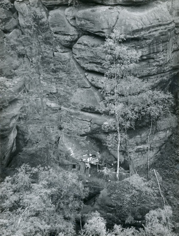 Das Bild ist eine Schwarz-weiß-Fotografie der sogenannten Fernblick-Boofe im Kreis Sebnitz aus einiger Entfernung. Zu sehen ist vor allem eine massive Felswand, die von Spalten durchzogen und teilweise mit Bäumen bewachsen ist. Im unteren Drittel des Bildes ist an den Fels angebaut die Holzverkleidung der Boofe erkennbar. 