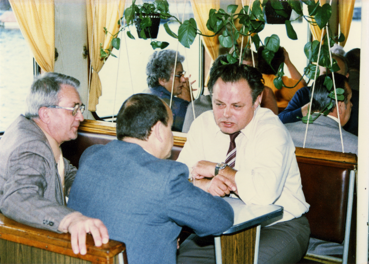 Das farbige Lichtbild zeigt im Vordergrund drei Herren, die sich an einem Tisch gegenüber sitzen und in ein Gespräch vertieft sind. Im Hintergrund sitzen weitere durchmischte Personengruppen an Tischen zusammen, es handelt sich sich um eine Bootsfahrt.