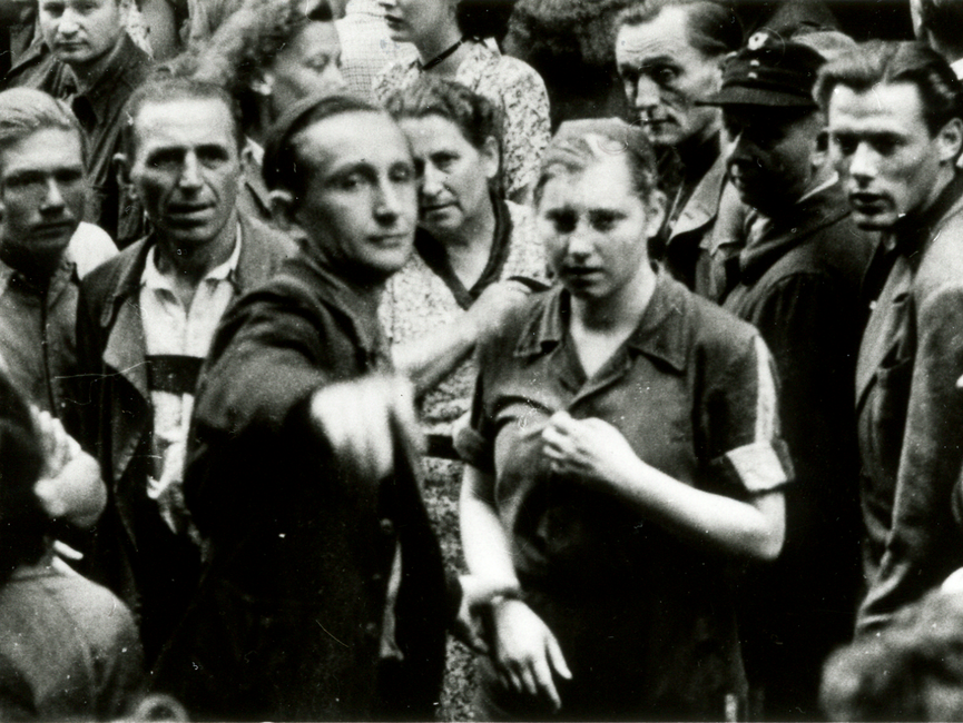 ie Schwarz-Weiß-Aufnahme zeigt eine Gruppe von Menschen. In der Mitte stehen ein Mann und eine Frau. Der Mann zeigt in Richtung des Betrachters oder der Betrachterin des Bildes.