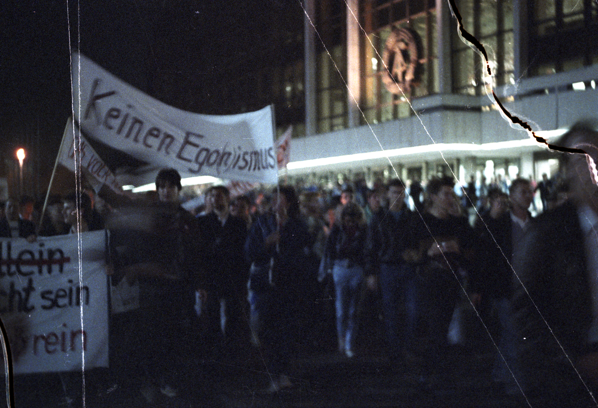 Ein Demonstrationszug vor dem Palast der Republik in Berlin-Mitte am Abend des 24. Oktober 1989 aus Protest gegen die Ernennung von Egon Krenz zum neuen Staatschef der DDR. Die Menschen marschieren am späten Abend, es ist dunkel und sie halten Kerzen in der Hand. Im Hintergrund ist der Palast der Republik zu sehen, das Staatswappen der DDR mit Hammer, Zirkel und Ährenkranz über dem Eingang hell erleuchtet. Die Demonstranten tragen Transparente, eines davon ist lesbar. "Keinen Ego(n)ismus" steht darauf.