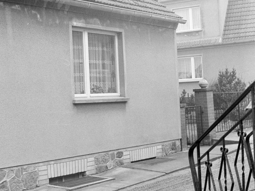Das schwarz-weiße Lichtbild zeigt eine Hausecke eines Einfamilienhauses, der Haussockel ist mir Natursteinen gefliest. Zu sehen ist eines der Fenster sowie der anschließende Zaun des Nachbargrundstückes. In der rechten Bildecke sieht man das schmiedeeiserne Geländer einer Treppe.