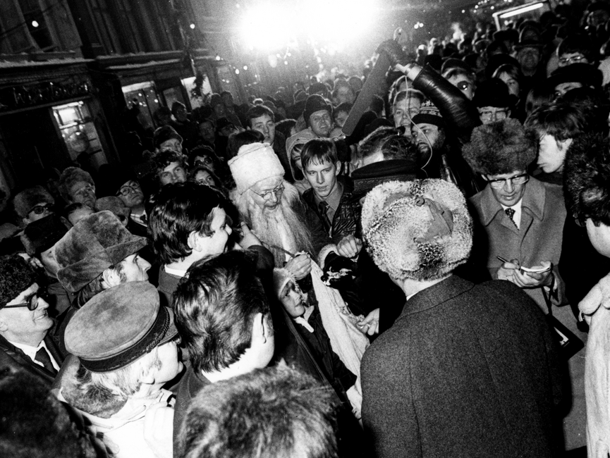 Der Ausschnitt der Schwarzweißaufnahme zeigt Helmut Schmidt und Erich Honecker (von hinten fotografiert) auf dem Güstrower Weihnachtsmarkt. Es hat sich eine große Menschenmenge um die beiden Staatsmänner versammelt. Die Menschenansammlung zieht sich weit bis in die Straße hinein. Vor Erich Honecker und Helmut Schmidt ist auch ein Mann - verkleidet als Weihnachtsmann - zu erkennen. Ein Mann hinter Helmut Schmidt scheint auf einem kleinen Block in der Hand Notizen zu schreiben. Ein anderer Mann hält von hinten ein Mikrofon in Richtung Helmut Schmidt und Erich Honecker hin. Ein Mann unten links im Bild trägt die für Helmut Schmidt typische Schirmmütze. 