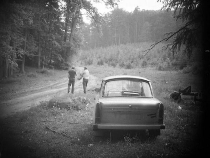 Das schwarz-weiße Lichtbild zeigt einen Waldrand. Auf einem losen Pfad sind zwei Männer von hinten zu erkennen. Im Bildfokus steht das Heck eines Trabant. Die Ecken sind abgedunkelt, vermutlich wurde das Foto verdeckt aufgenommen.