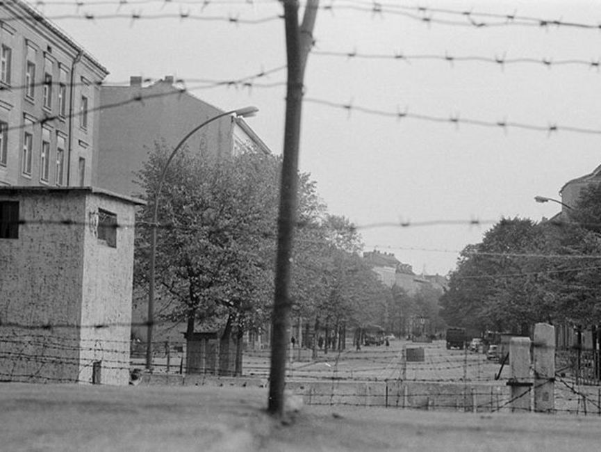 Auf dem schwarz-weißen Lichtbild wurde eine Straße hinter dem Stacheldraht auf der Berliner Mauer fotografiert. Die Straße zwischen den Häuserzeilen wird von Bäumen eingefasst, von den Grenzsicherungsanlagen ist kaum etwas zu erkennen.